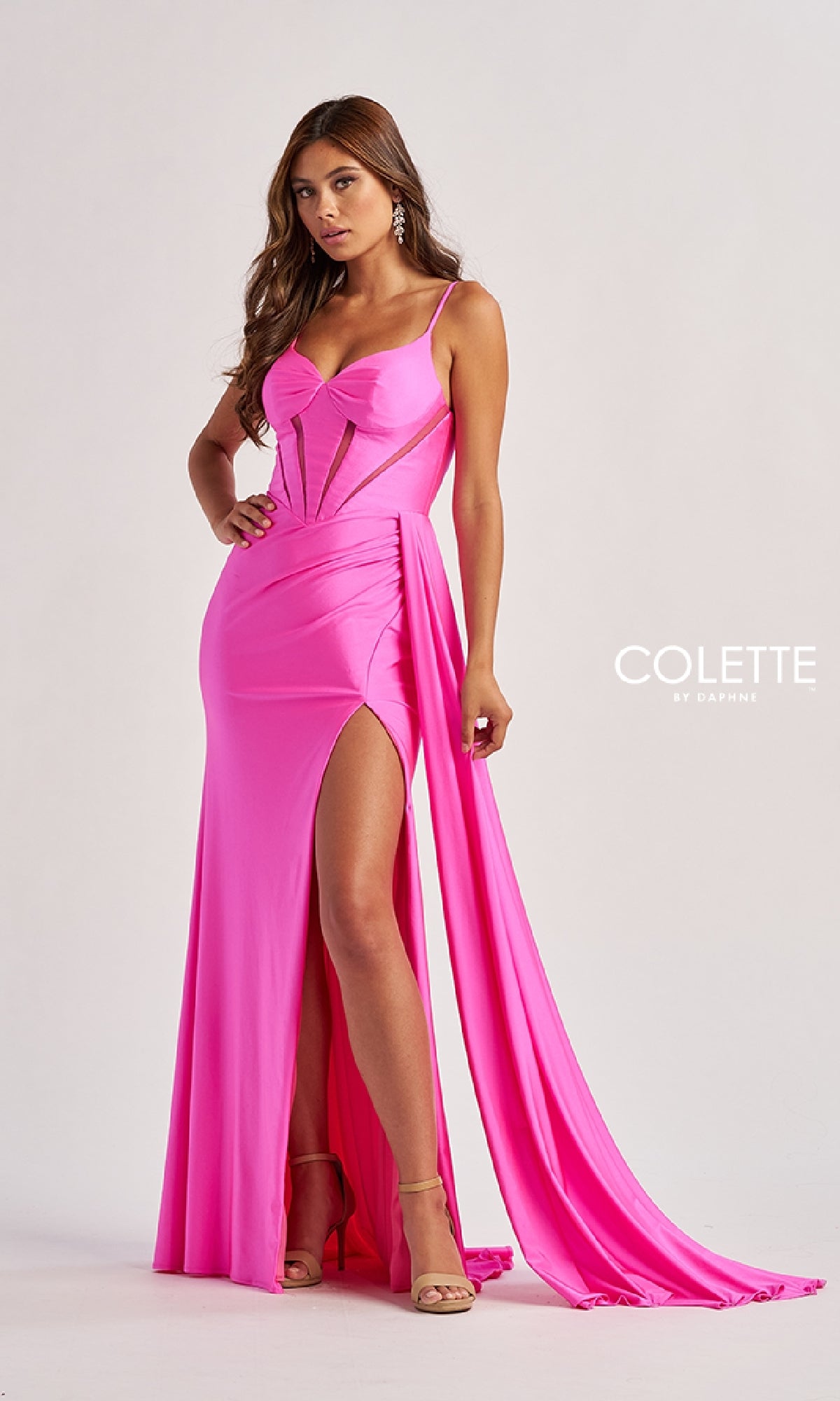 A woman stands against a plain background wearing the Sheer-Cut-Out Long Prom Dress: Colette CL8480—a bright pink, floor-length dress featuring a thigh-high slit, lace-up fitted bodice, and spaghetti straps.