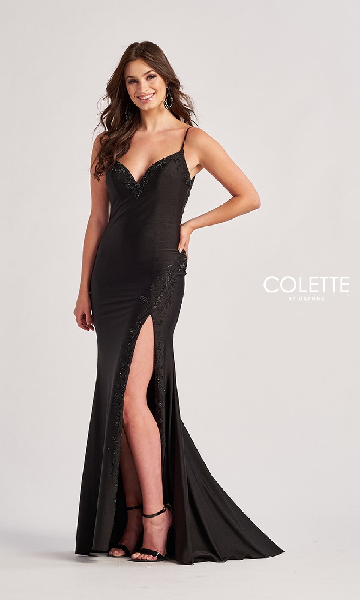 A woman models the Colette CL8485 Long Prom Dress—a fitted black evening gown with a thigh-high slit, corset bodice, thin straps, and subtle embellishments—standing against a plain background.
