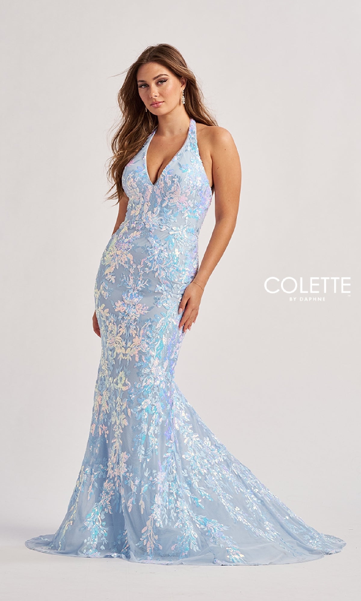 A woman stands against a plain background wearing the Colette CL8495 Long Prom Dress—a light blue floral lace halter gown with a fitted silhouette, halter v-neckline, and slight train.
