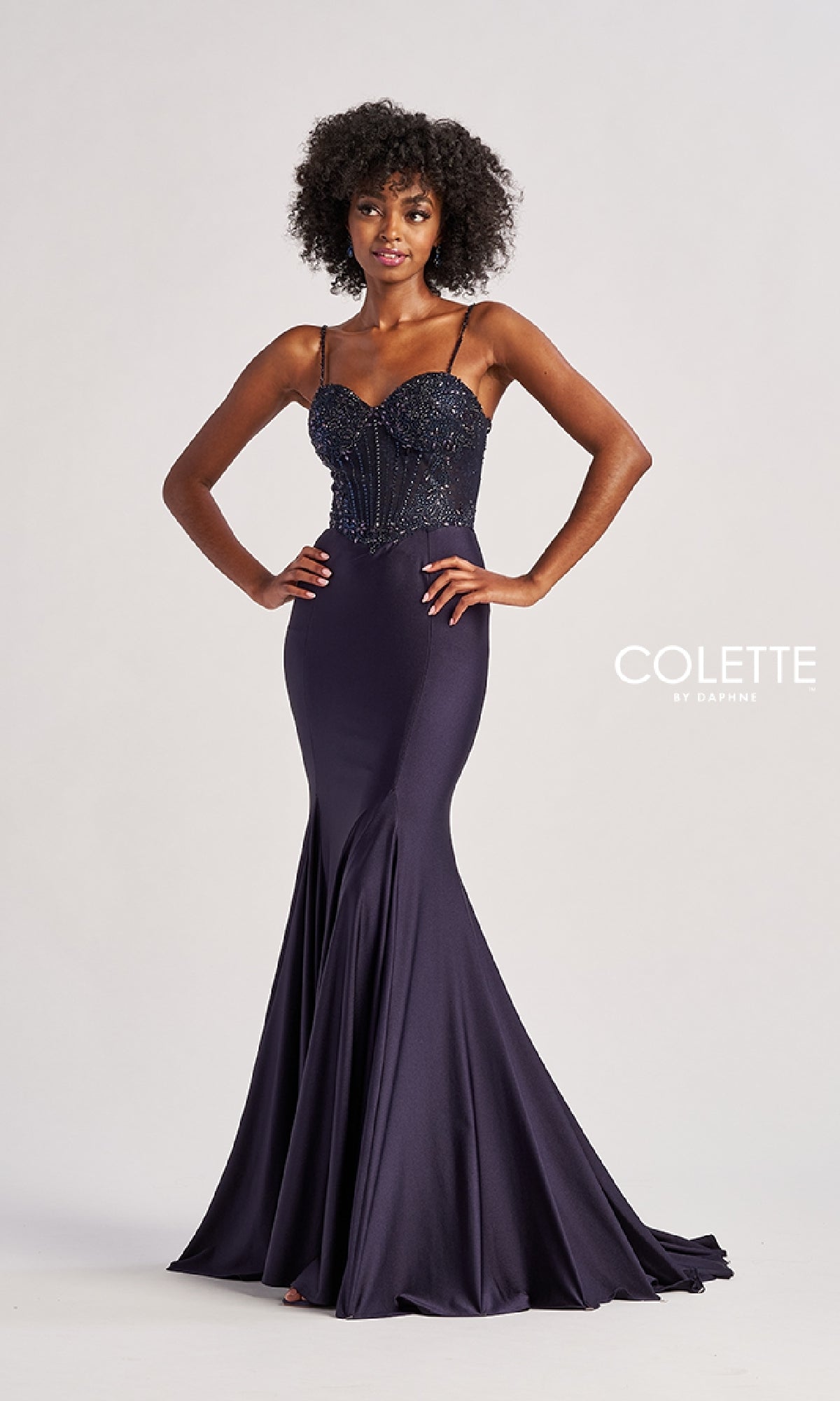 A woman in a navy blue, floor-length mermaid prom dress with a beaded lace corset bodice and thin straps—Colette CL8515—stands hands on hips against a plain background, with the 