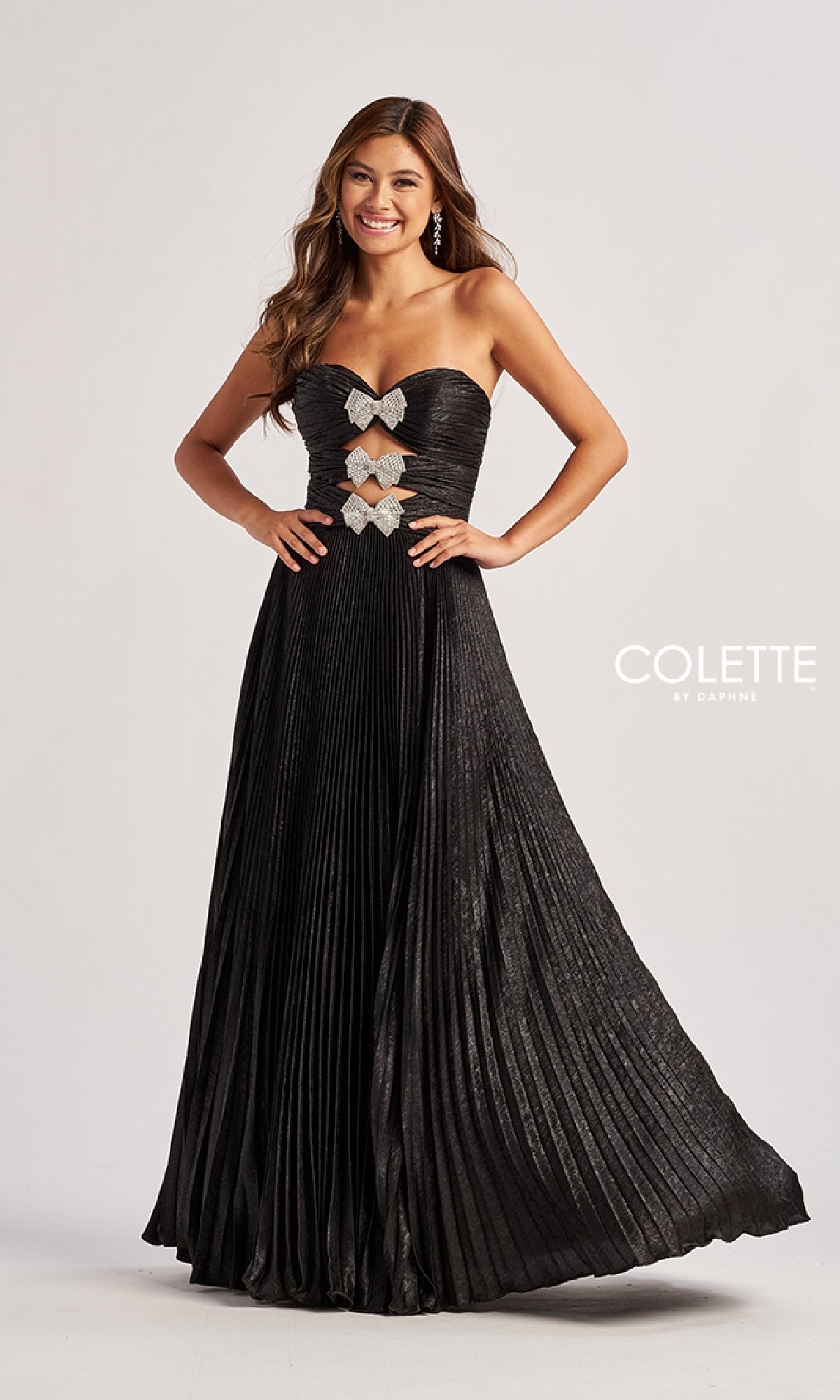 A woman stands against a plain background in the Colette CL8520 strapless pleated long prom dress featuring silver butterfly accents and rhinestone bows on the bodice, with the 