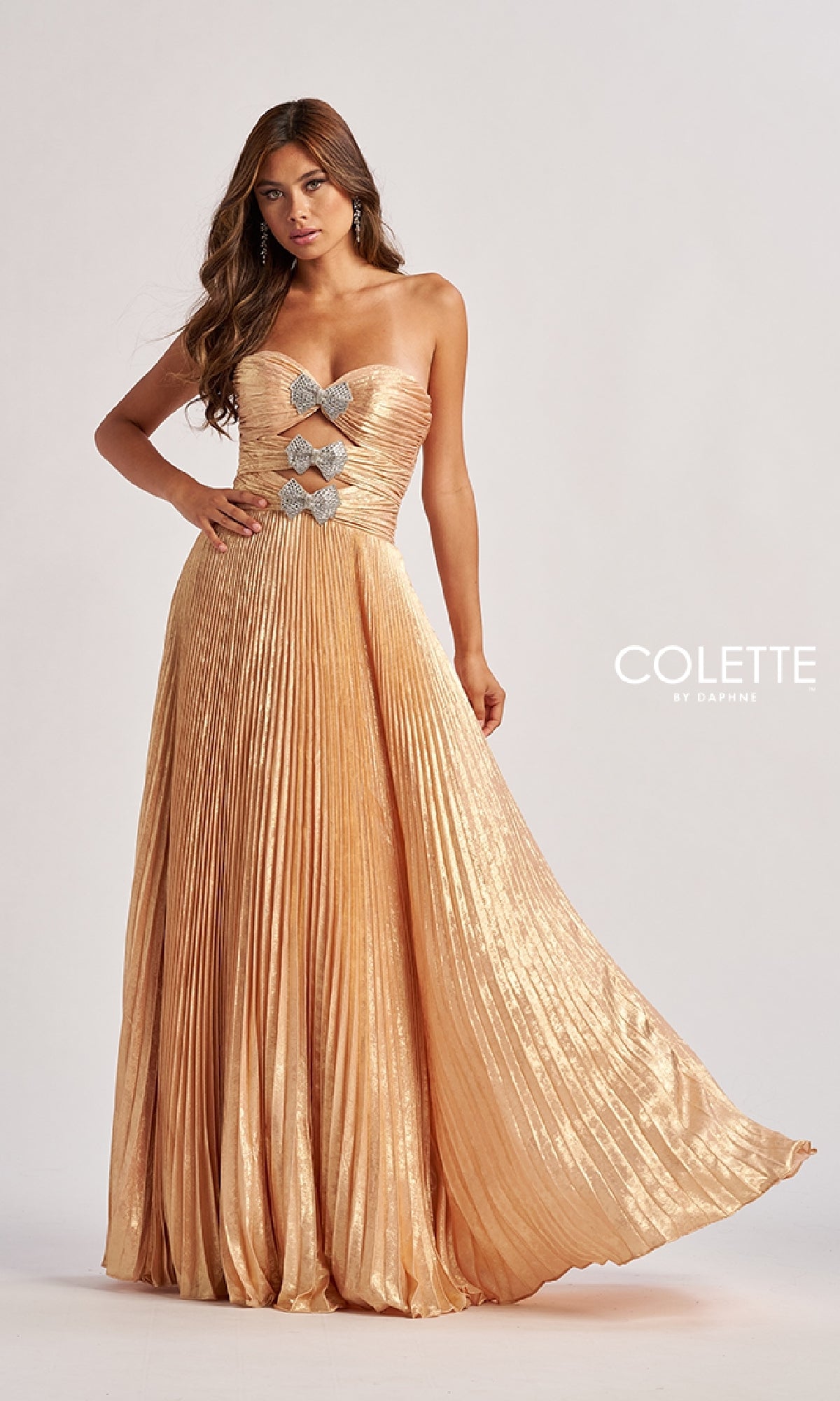 A woman wears the Colette CL8520 Strapless Pleated Long Prom Dress in metallic gold with rhinestone bow accents on the bodice, standing against a plain background. 