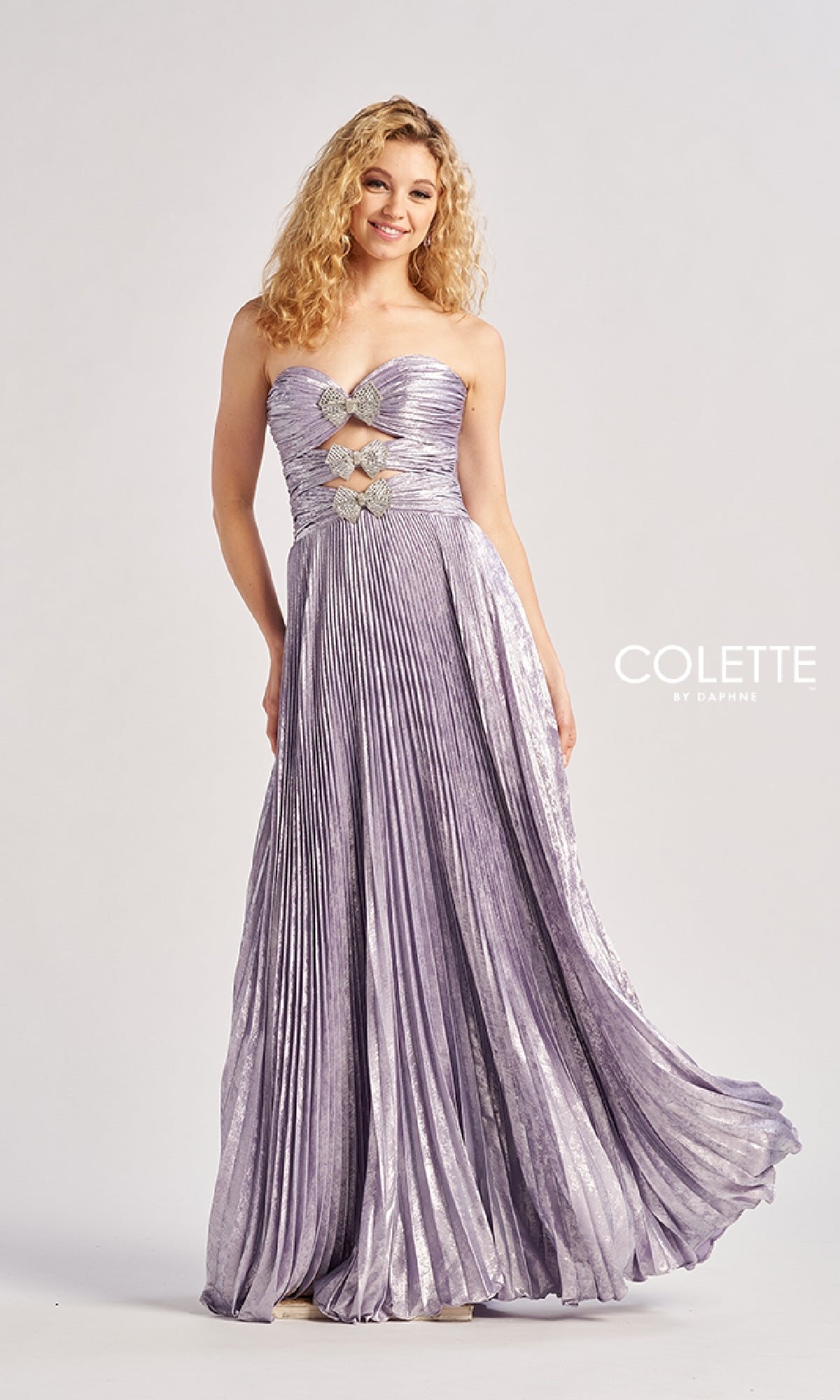 A woman models the Colette CL8520 Strapless Pleated Long Prom Dress in metallic lavender with geometric silver embellishments and rhinestone bows on the bodice, set against a plain background.