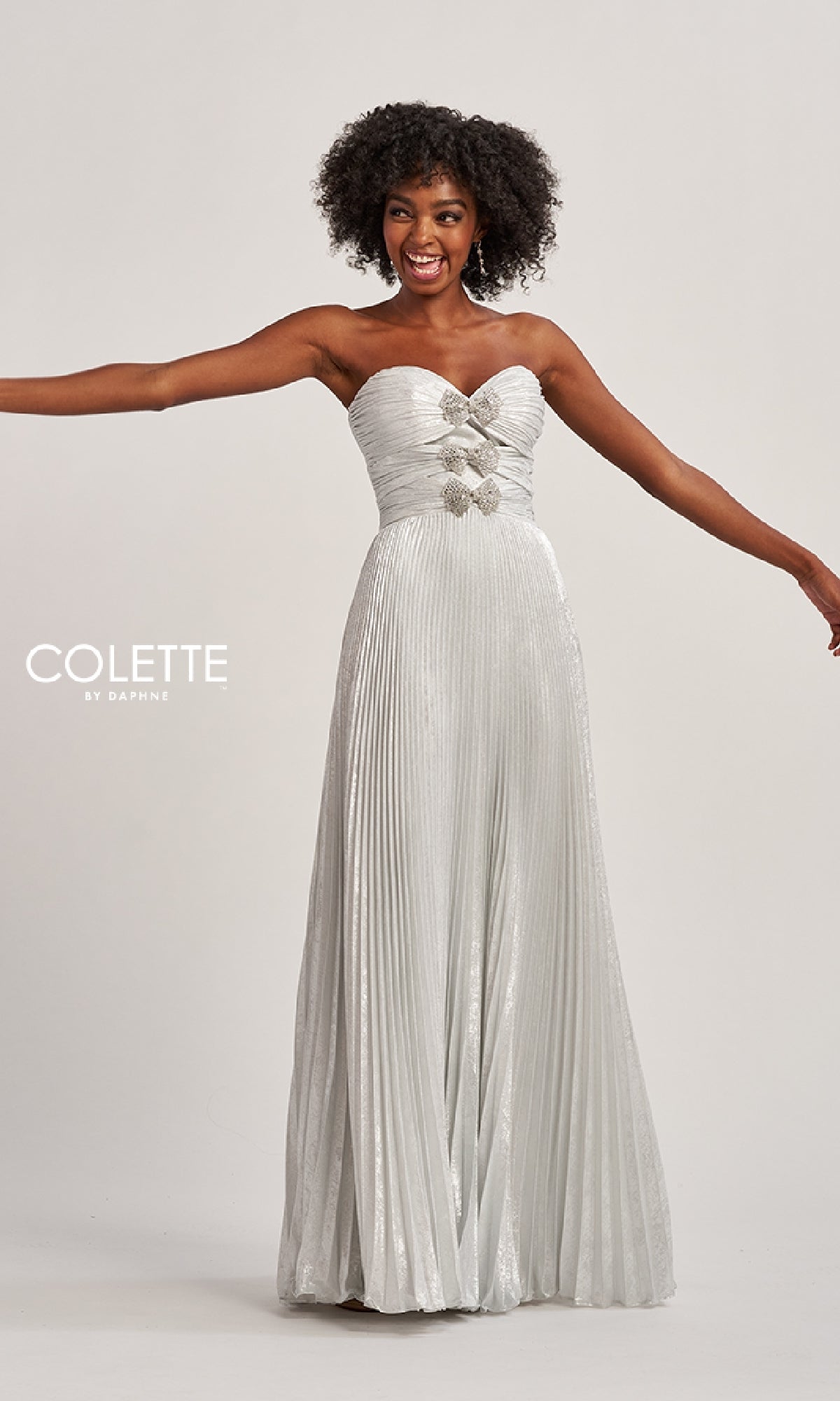Woman modeling the Colette CL8520, a strapless long pleated prom dress with rhinestone bows at the bust, standing with arms outstretched against a plain background.