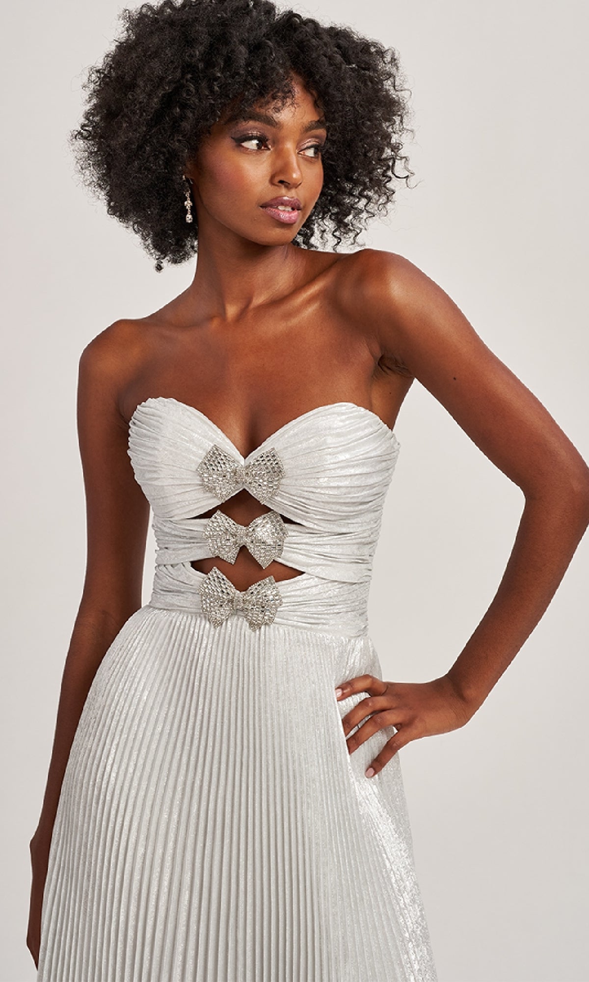 A woman stands against a plain background in the Colette CL8520, a strapless silver pleated A-line evening gown with rhinestone bows and bodice cutout details.