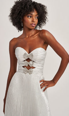A woman stands against a plain background in the Colette CL8520, a strapless silver pleated A-line evening gown with rhinestone bows and bodice cutout details.