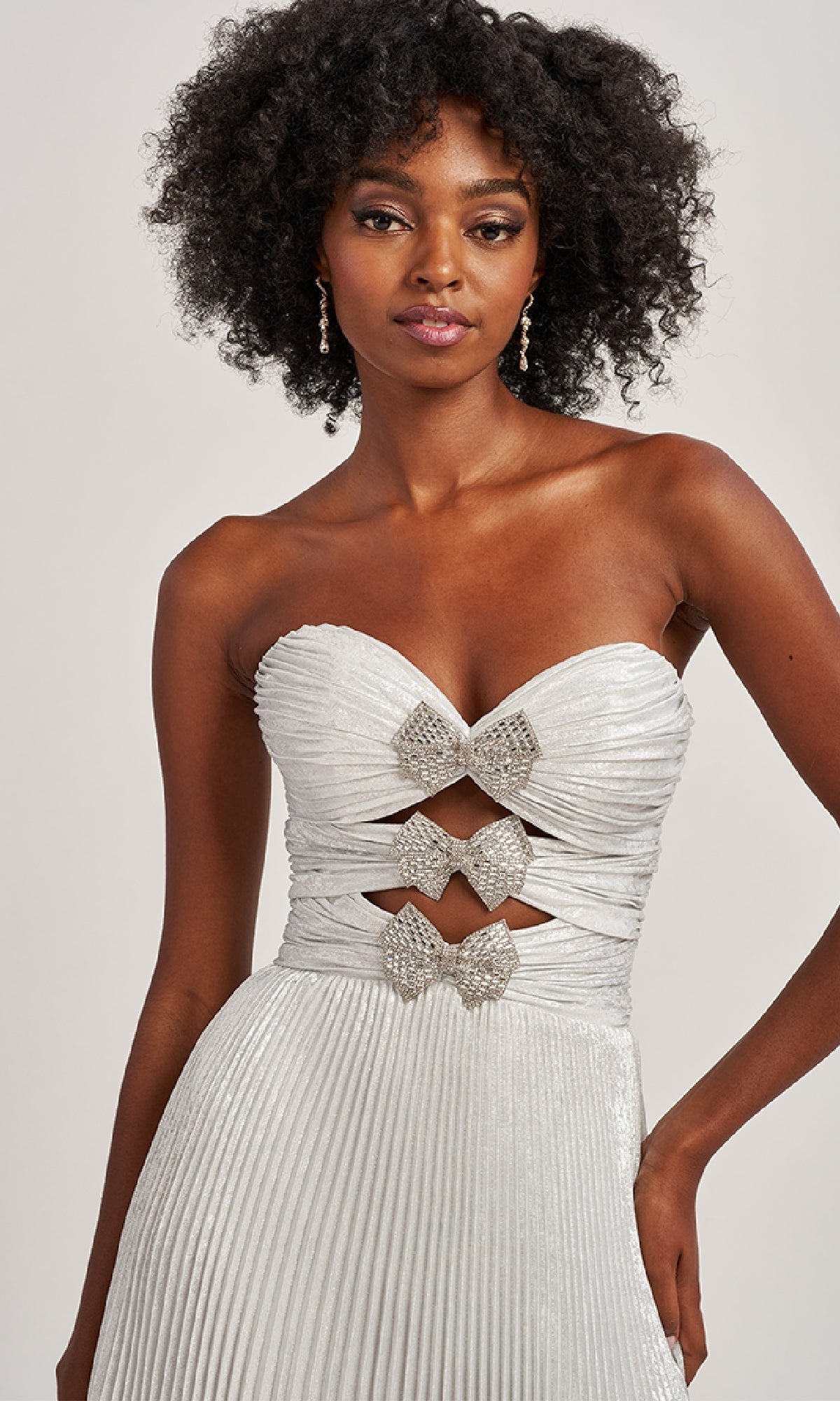 A woman with curly hair stands against a plain background wearing the Colette CL8520 strapless pleated long prom dress, which features three front cutouts each accented with sparkling rhinestone bows.