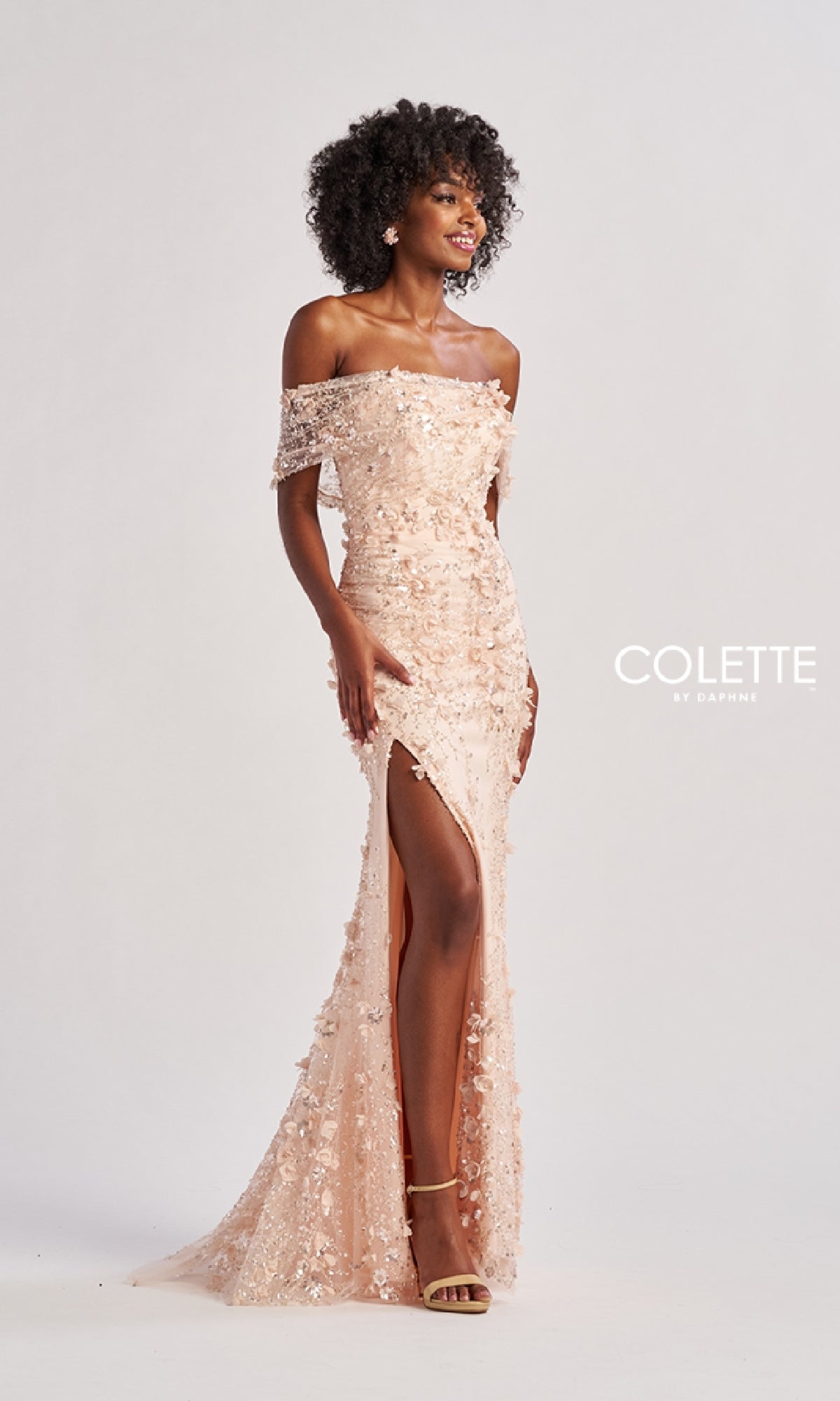 A woman stands smiling in front of a plain background, wearing the blush-colored Colette CL8530 long prom dress featuring an off-the-shoulder design, sequins, and a thigh-high slit. The 