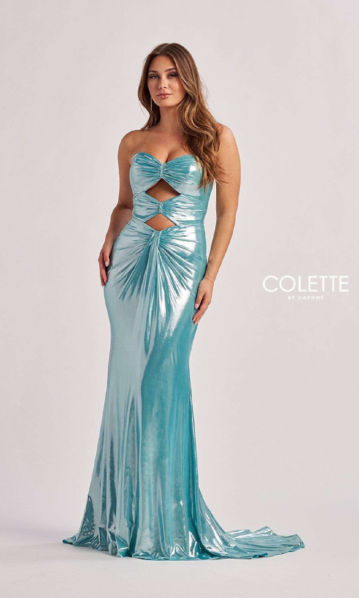 A woman wears the Long Prom Dress: Colette CL8545, a shiny teal, strapless gown with cut-out details and metallic finish. The fitted, floor-length dress stands out against a light background. 