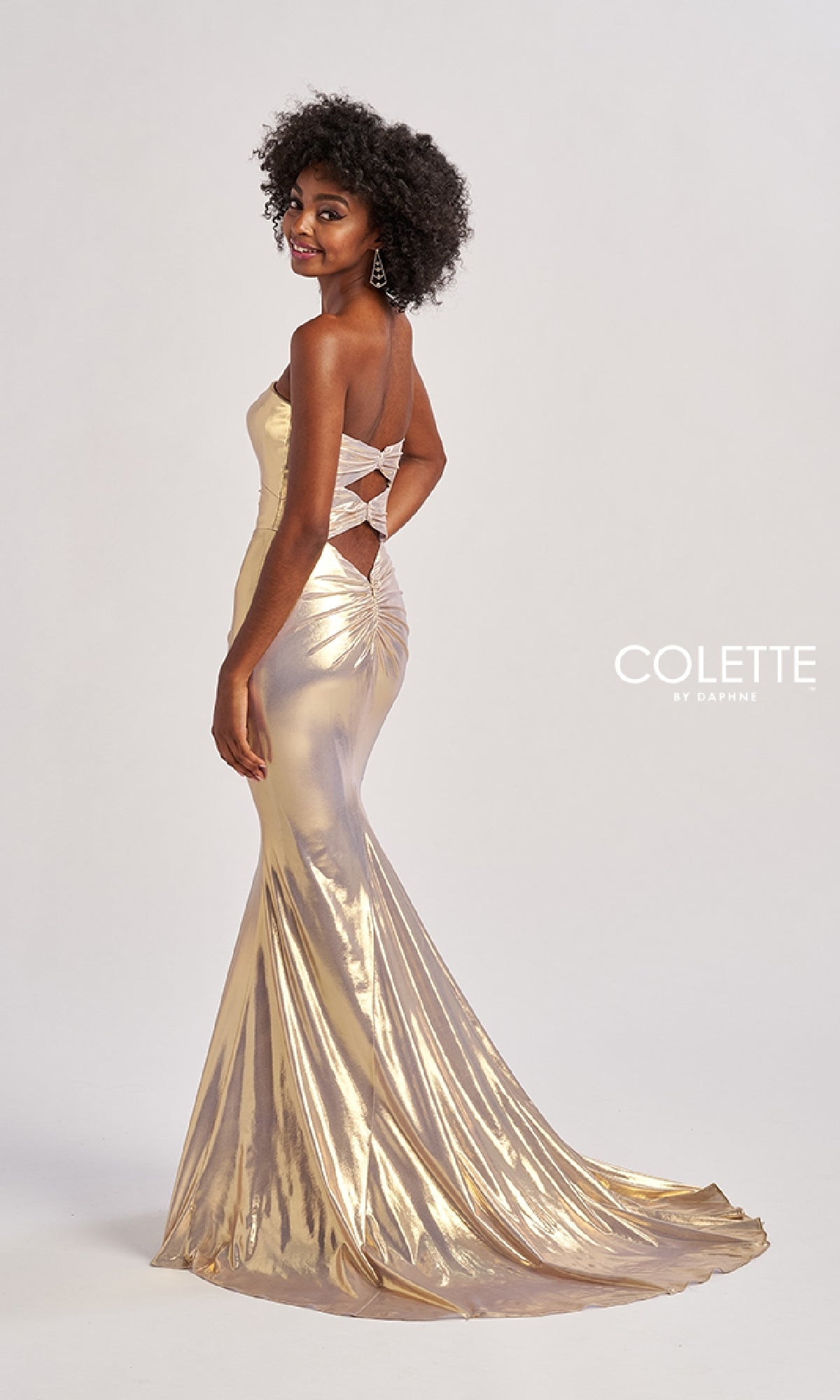 A woman wears the Colette CL8545 long prom dress in gold metallic, featuring a fitted floor-length silhouette and an open crisscross back, standing against a plain background.
