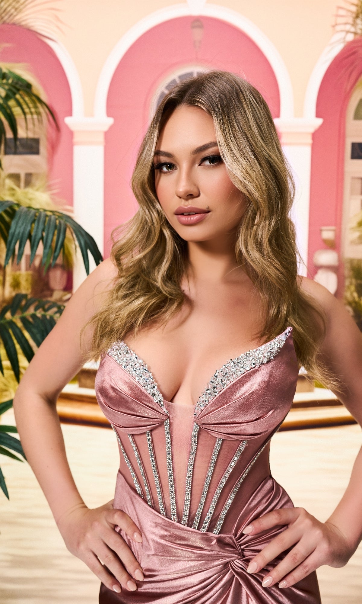 A woman with long blonde hair wears the Sheer-Bodice Long Prom Dress Colette CL8560, a pink strapless gown with a corset bodice, standing in front of a pink building with arched windows and palm leaves.