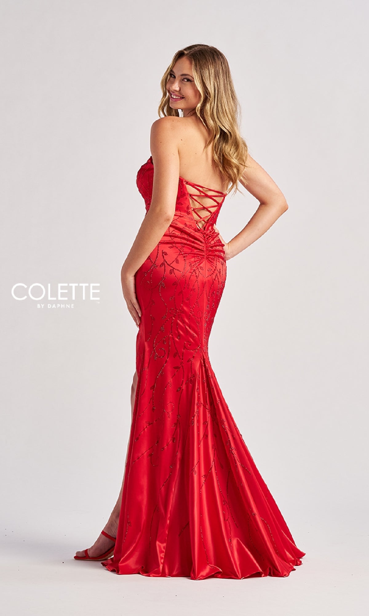 A woman models the Colette CL8570 long prom dress, featuring a strapless design, lace-up strappy open back, and beaded sequin appliques, posing elegantly against a plain background.