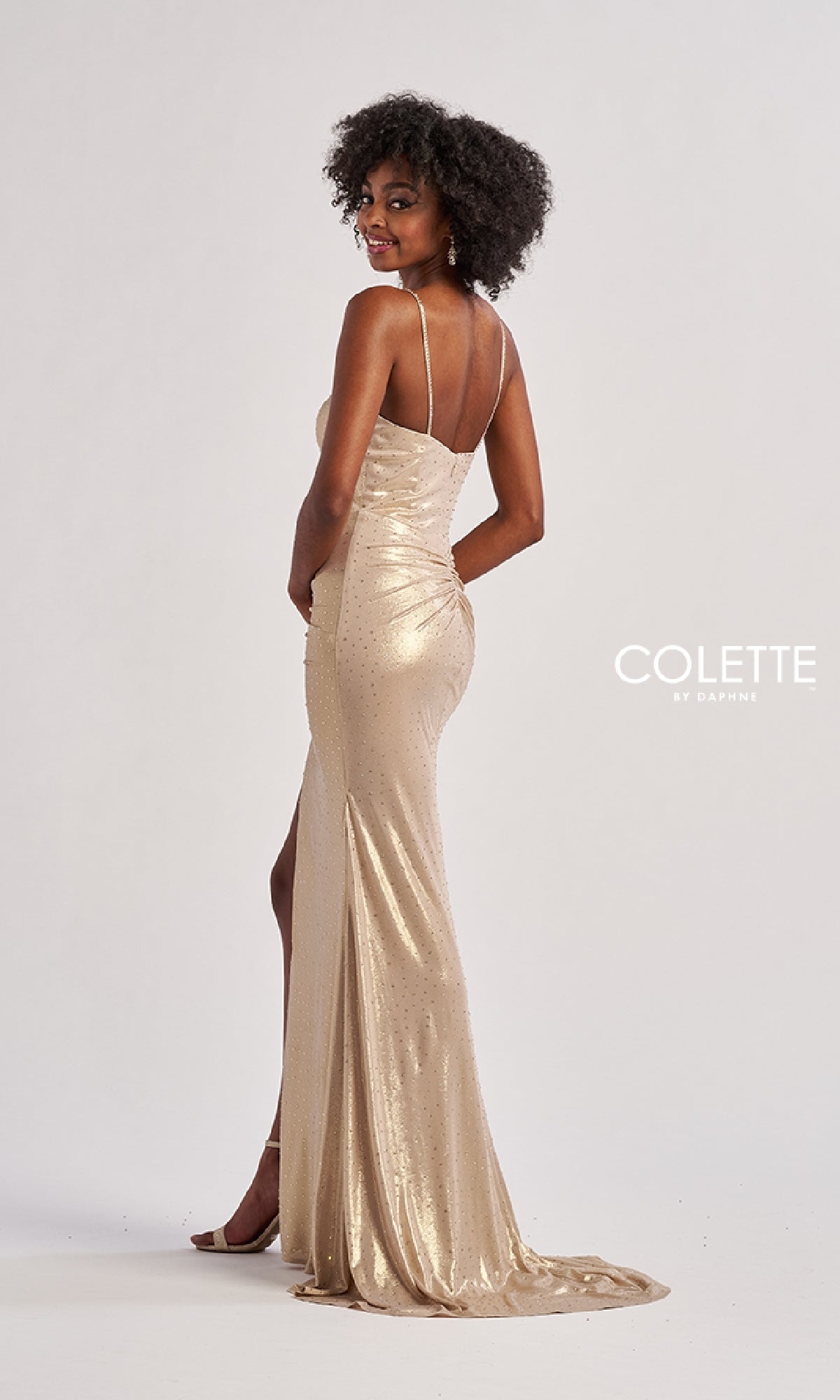 A woman poses against a plain white background wearing the Long Prom Dress: Colette CL8575, a metallic stretch jersey gown with stone accents, thin straps, and a thigh-high side slit.