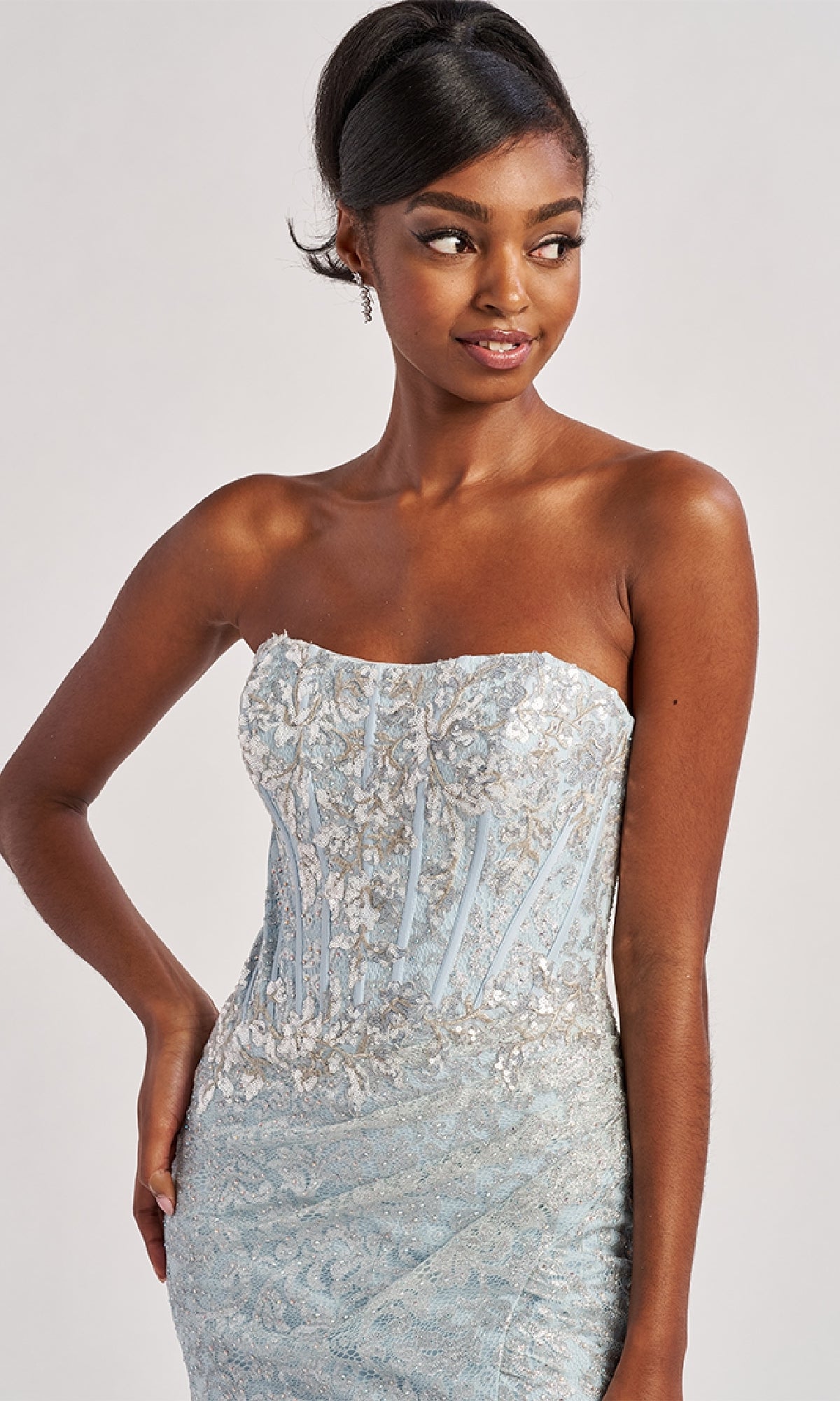 A woman poses with one hand on her hip, wearing the Colette CL8595 Long Prom Dress—a strapless light blue gown with silver embroidery—and looks to the side.