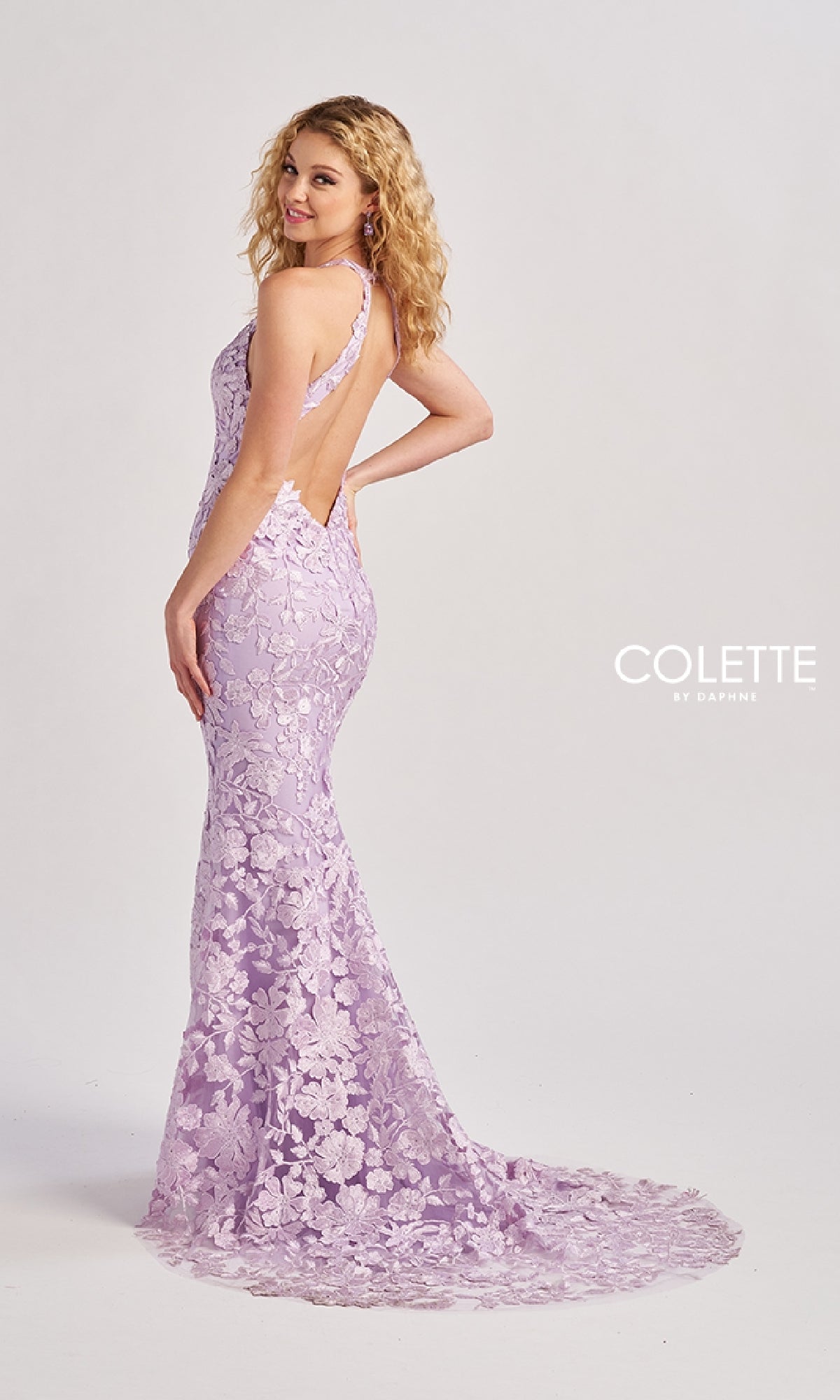 A woman poses sideways, hand on hip, in the Colette CL8665 long prom dress—a purple mermaid lace gown with an open back and a long, floral-patterned train—set against a plain background.