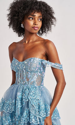 A woman models the Colette CL8690 long prom dress featuring an off-shoulder fitted bodice, sheer accents, and a blue sequin ruffle-tiered A-line skirt, posing against a plain backdrop.