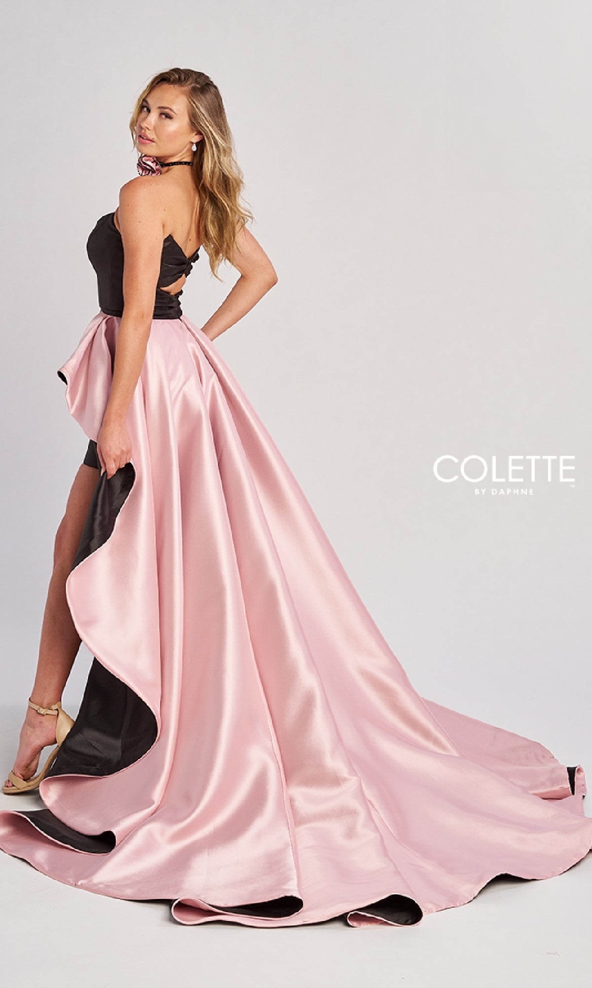 A woman poses in the Colette CL8695 long prom dress—a strapless black and pink high-low gown with a dramatic satin overskirt train—paired with strappy heels.