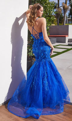 A woman wearing the Lace-Up Long Mermaid Shimmer Prom Dress CM328 in bright blue stands outdoors by a white wall and greenery.