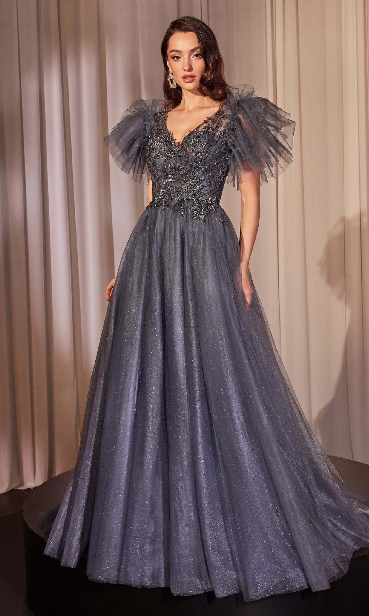 A woman stands before draped curtains wearing the Ladivine CM359 Flutter-Sleeve Blue Prom Ball Gown, featuring a long glittery skirt, fitted bodice, floral details, and dramatic tulle flutter sleeves.