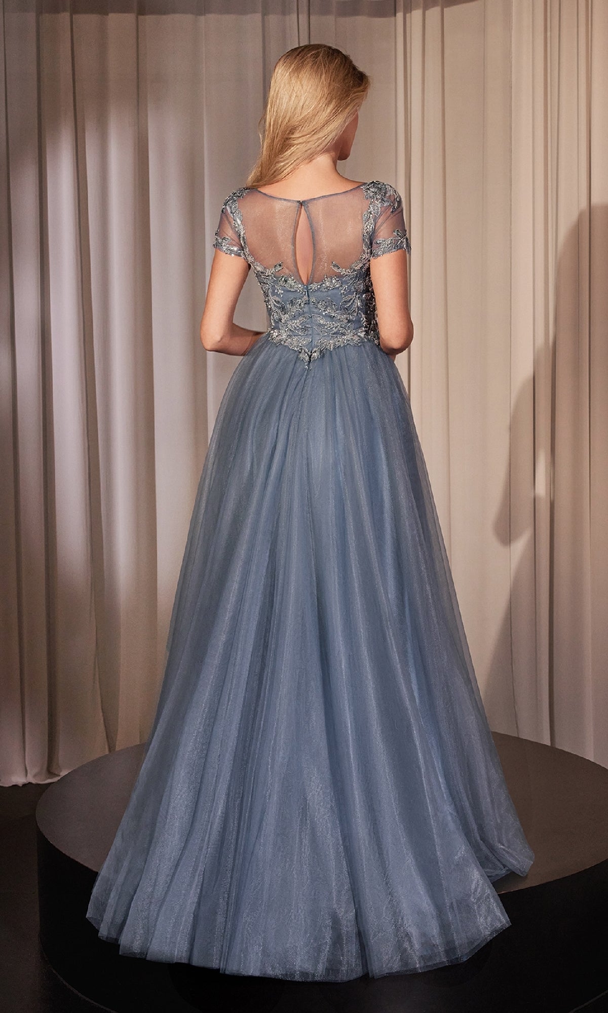 A woman stands on a platform facing away, wearing the Short-Sleeve Blue Prom Ball Gown: Ladivine CM360. The dress features sheer short sleeves, an embroidered beaded sweetheart bodice, and a flowing A-line skirt. Beige curtains are in the background.