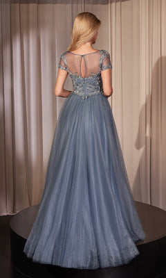 A woman stands on a platform facing away, wearing the Short-Sleeve Blue Prom Ball Gown: Ladivine CM360. The dress features sheer short sleeves, an embroidered beaded sweetheart bodice, and a flowing A-line skirt. Beige curtains are in the background.