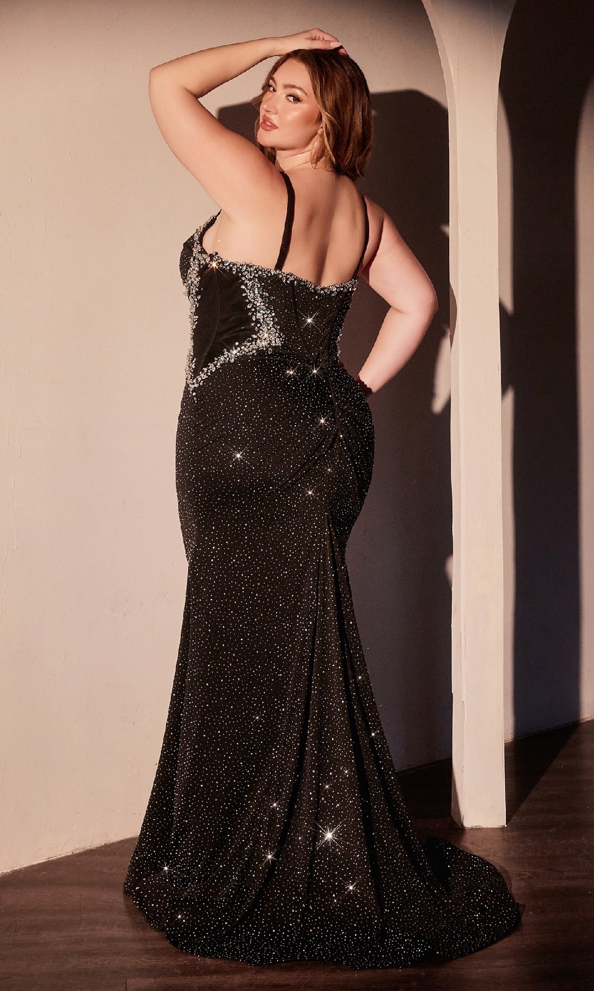 A woman poses with her back to the camera in the Ladivine CM365C, a plus-size long black prom dress with beaded silver detailing, standing in a softly lit room.