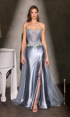 A woman in a Ladivine CM392 long prom dress in dusty blue, featuring a metallic strapless design with a sheer corset bodice, thigh-high slit, and detachable overskirt, stands near a white piano and a lit candle.