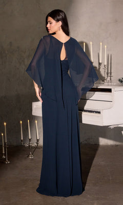 A woman wearing the Ladivine CR884 long formal dress, featuring dark blue chiffon and sheer embellished cape sleeves, stands facing away from the camera in front of a white piano and candelabras.
