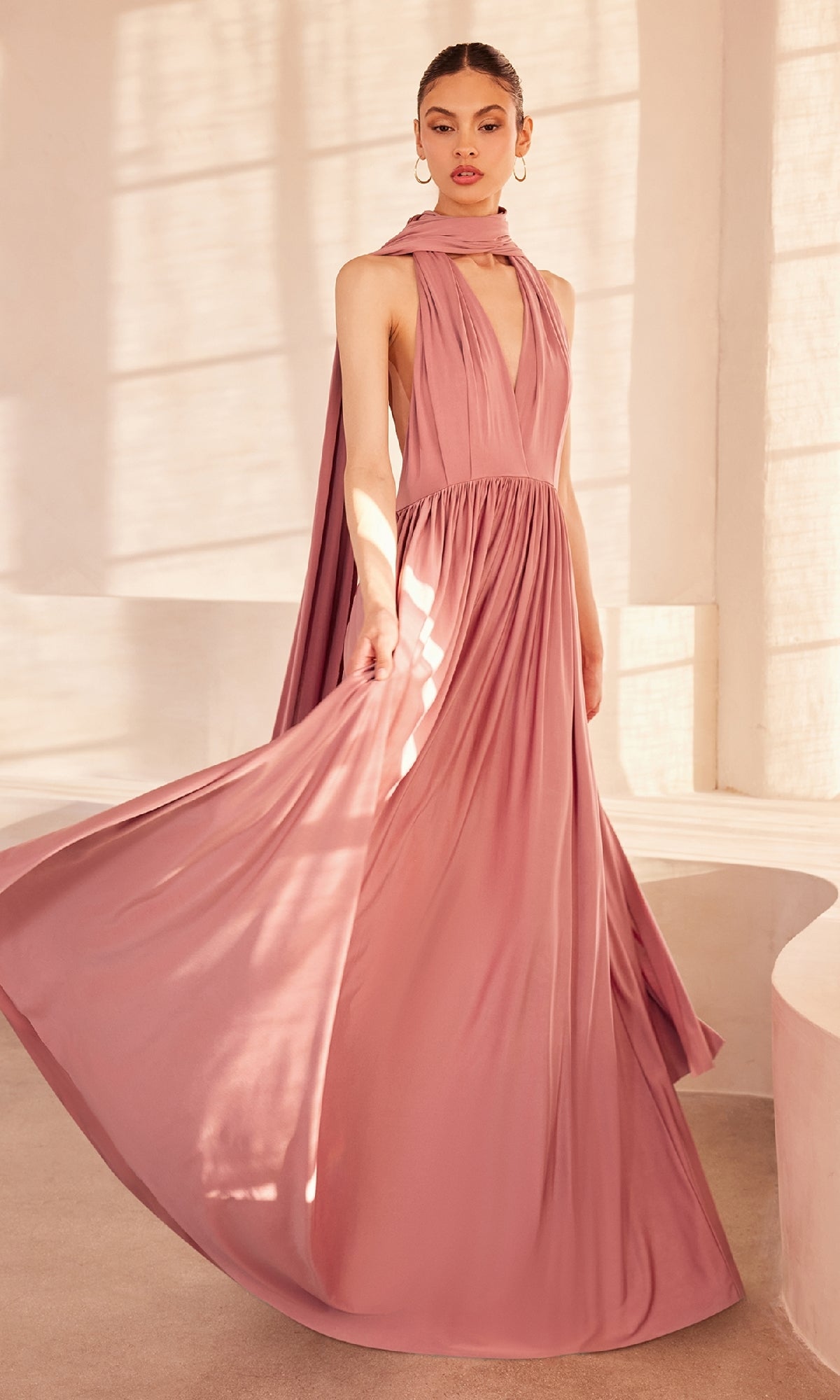 A woman stands in a sunlit room with light walls wearing the Amoris CV01 Multi-Wrap Long A-Line Prom Dress—a flowing, sleeveless mauve gown with a gathered waist and matching scarf.