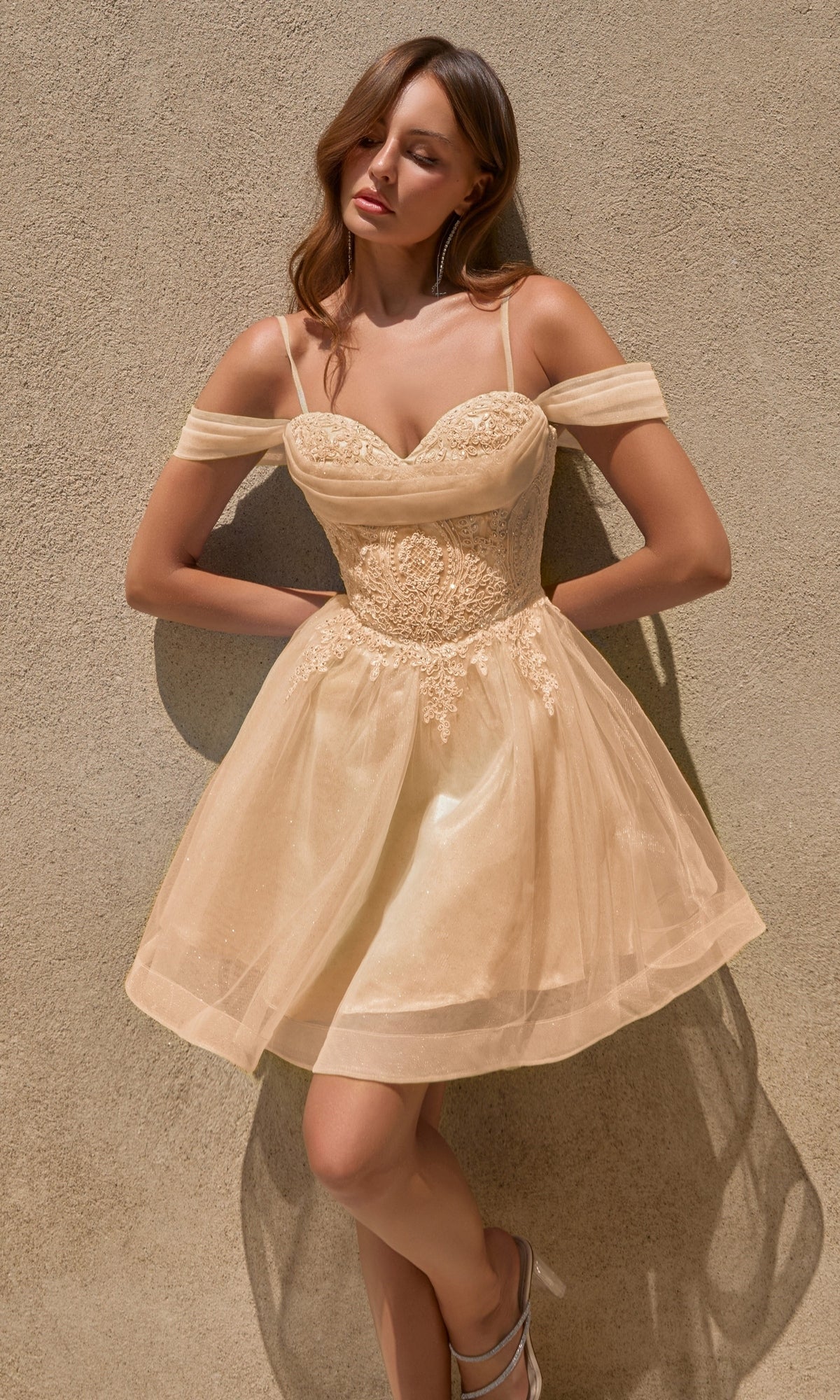 A woman in the Ladivine CY024 short homecoming dress in champagne gold, featuring off-shoulder styling and beaded lace applique details, stands against a textured tan wall, looking down with her arms resting at her sides.