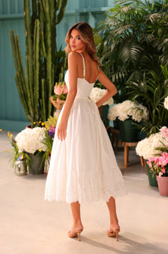 A woman wearing the White Casual Midi Dress: Celeste and nude heels stands indoors among potted plants and flowers, looking back over her shoulder.
