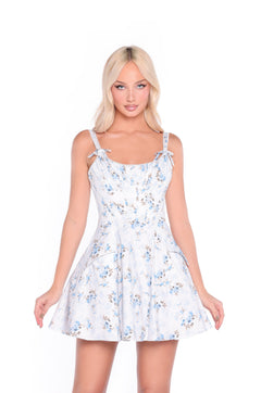 A woman stands against a white background wearing the Bluebell Floral Corset Summer Dress: Celestine, featuring light blue and white floral print, tie straps, and a flattering fit-and-flare silhouette.