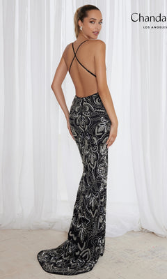 A woman stands in front of white curtains wearing the Sequin-Print Long Prom Dress: Chandalier 30087—a backless, floor-length black and silver gown with crisscross straps.
