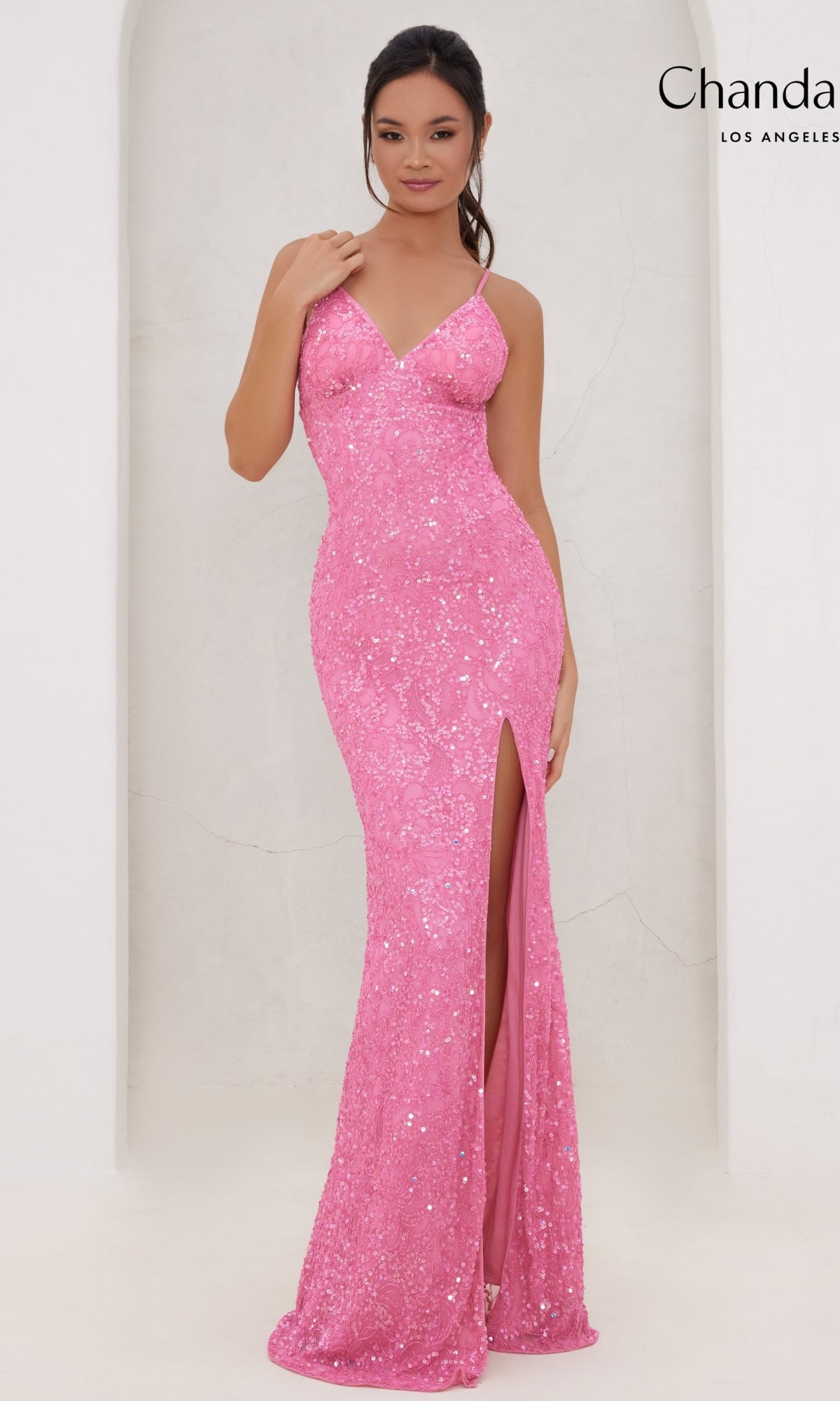 A woman wearing the Long Prom Dress 30118 by Chandalier—a fitted pink sequin gown with a v-neckline and thigh-high slit—stands against a light-colored background.