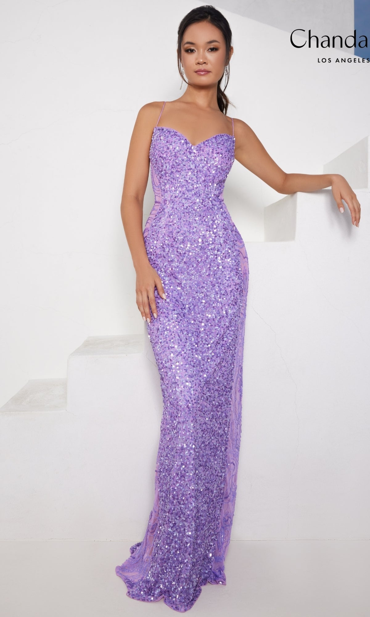 A woman stands near stairs against a white background, wearing the Long Prom Dress 30124 by Chandalier—a fitted, floor-length lavender sequin formal gown with thin straps.