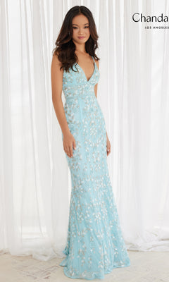 A woman stands before white curtains wearing the Long Prom Dress 30157 by Chandalier, a light blue, floor-length gown adorned with floral beads, silver accents, and featuring a deep V neckline.