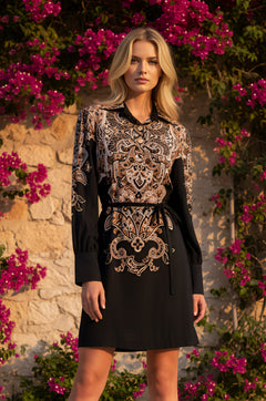 A woman stands outdoors in front of a stone wall with pink flowers, wearing the Clara Paisley-Print Short Casual Dress in Twilight Filigree, featuring long sleeves, an ornate print, and a matching belt.