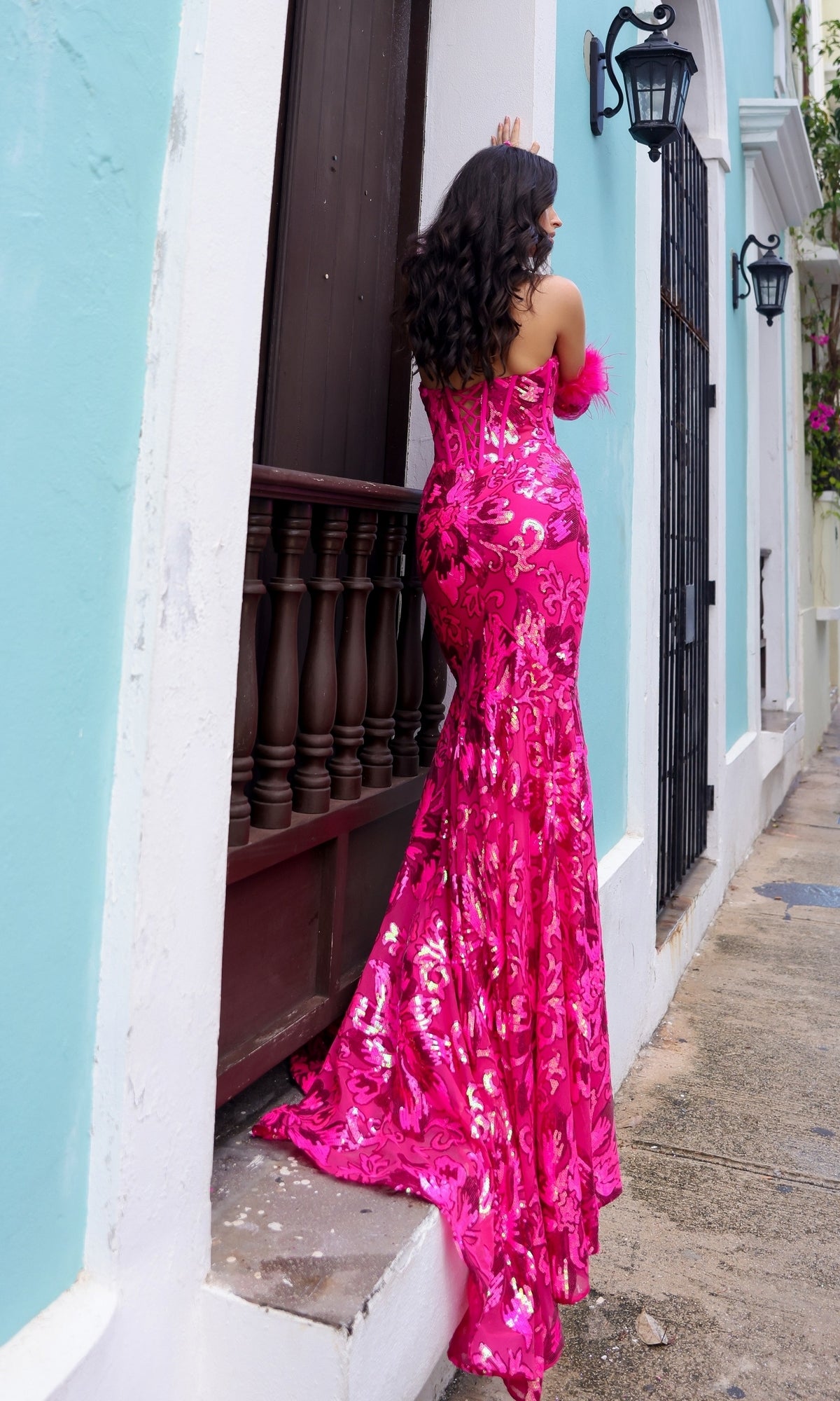 A woman in the Strapless Sequin-Print Prom Dress with Gloves D1269 stands on a narrow ledge outside a pastel blue building, facing away from the camera.