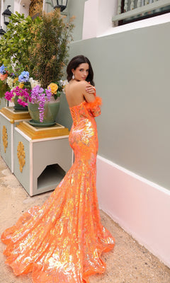 A woman stands outdoors in the Strapless Sequin-Print Prom Dress with Gloves D1269, a bright orange, fitted floor-length gown. She looks over her shoulder in elegance, floral arrangements nearby and feather-trimmed gloves completing her look.