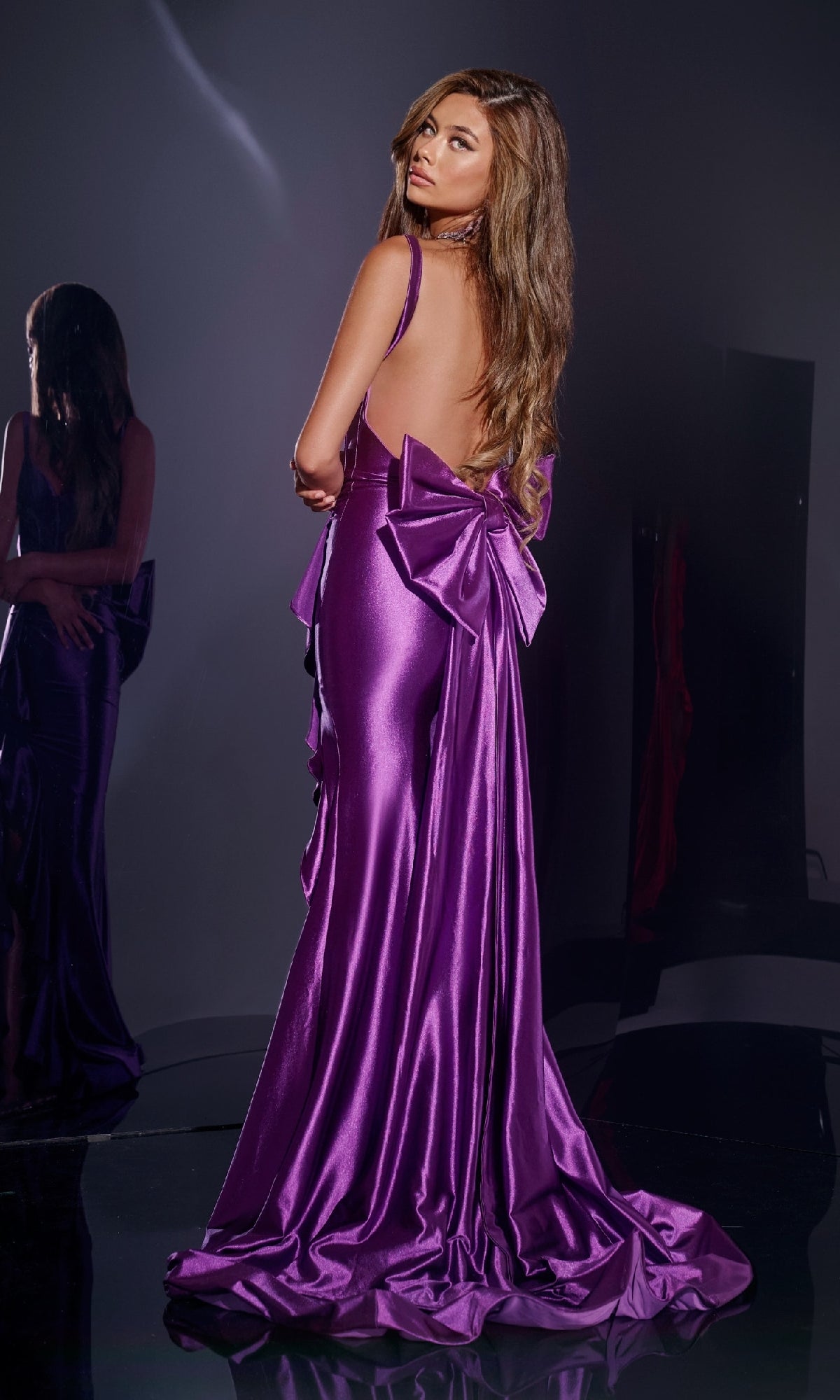 A woman in a shiny, floor-length, backless metallic prom dress with a corset bodice and large bow stands on a reflective surface, glancing over her shoulder. Her elegant Long Prom Dress: Jovani D5305 is mirrored behind her.