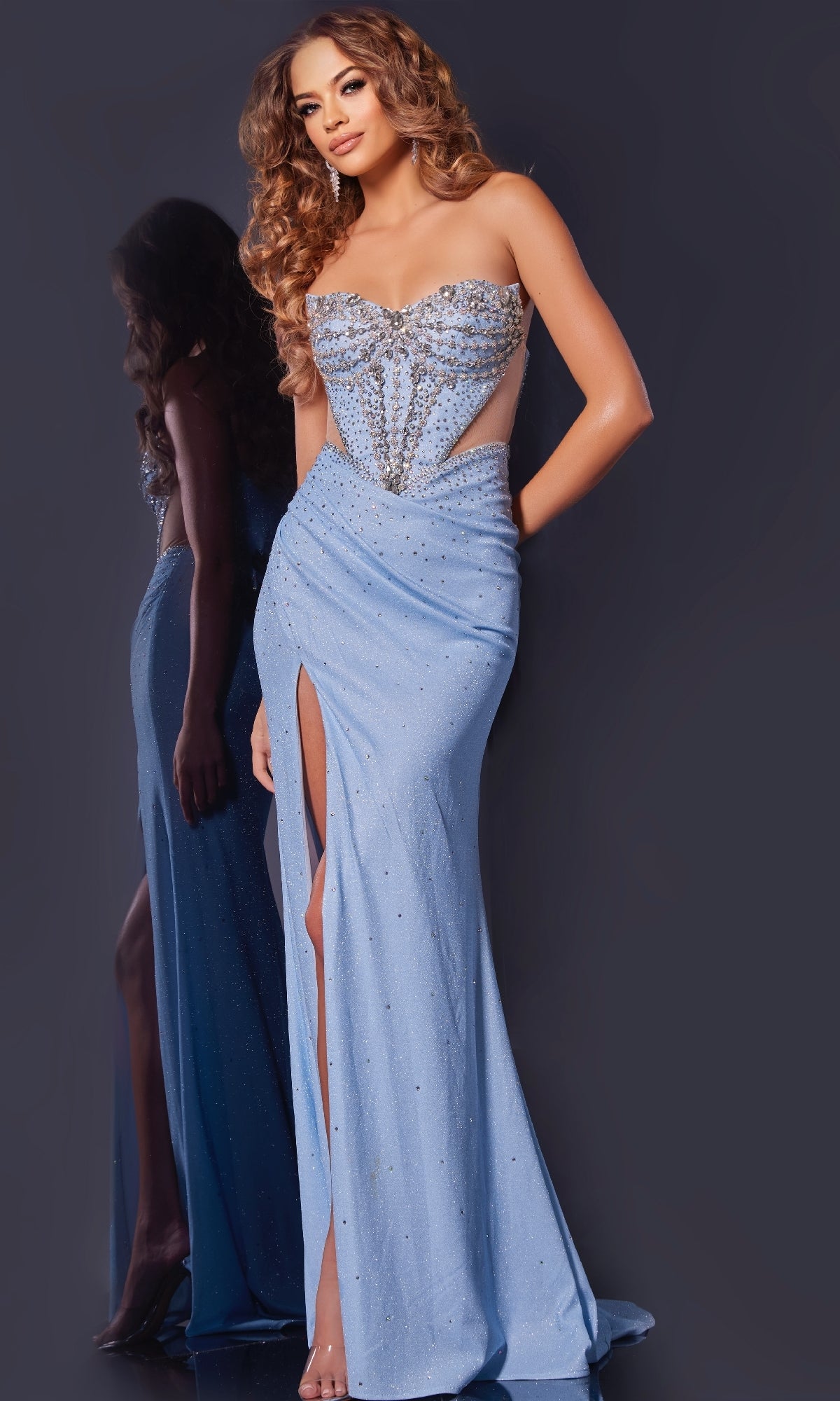 A woman in the Jovani D5414 long prom dress—strapless, blue, with a high slit, rhinestone details, and sheer side panels—stands against a dark background, highlighting the gown's elegance.