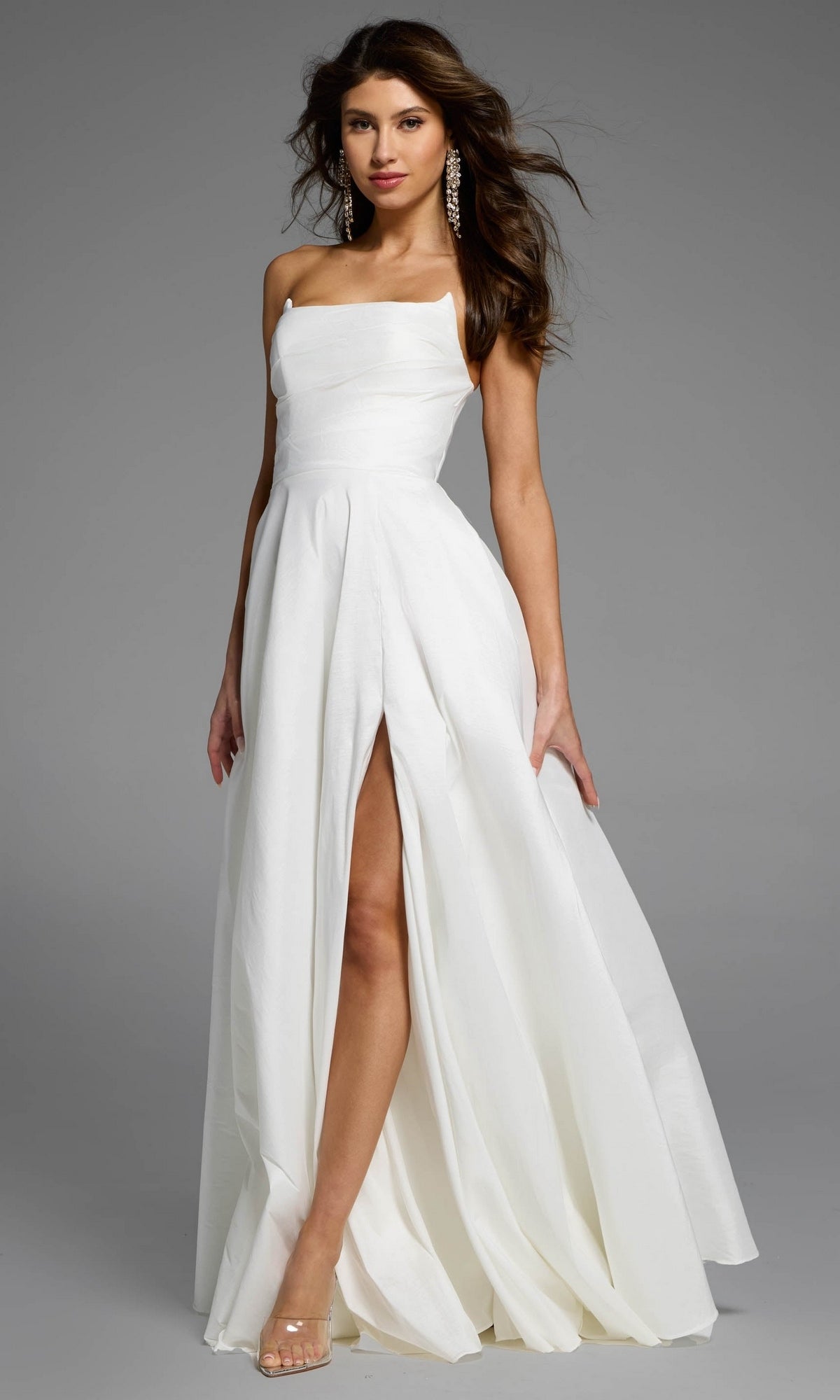 A model wears a strapless ivory Jovani D5822 prom dress with a thigh-high slit and clear heels.