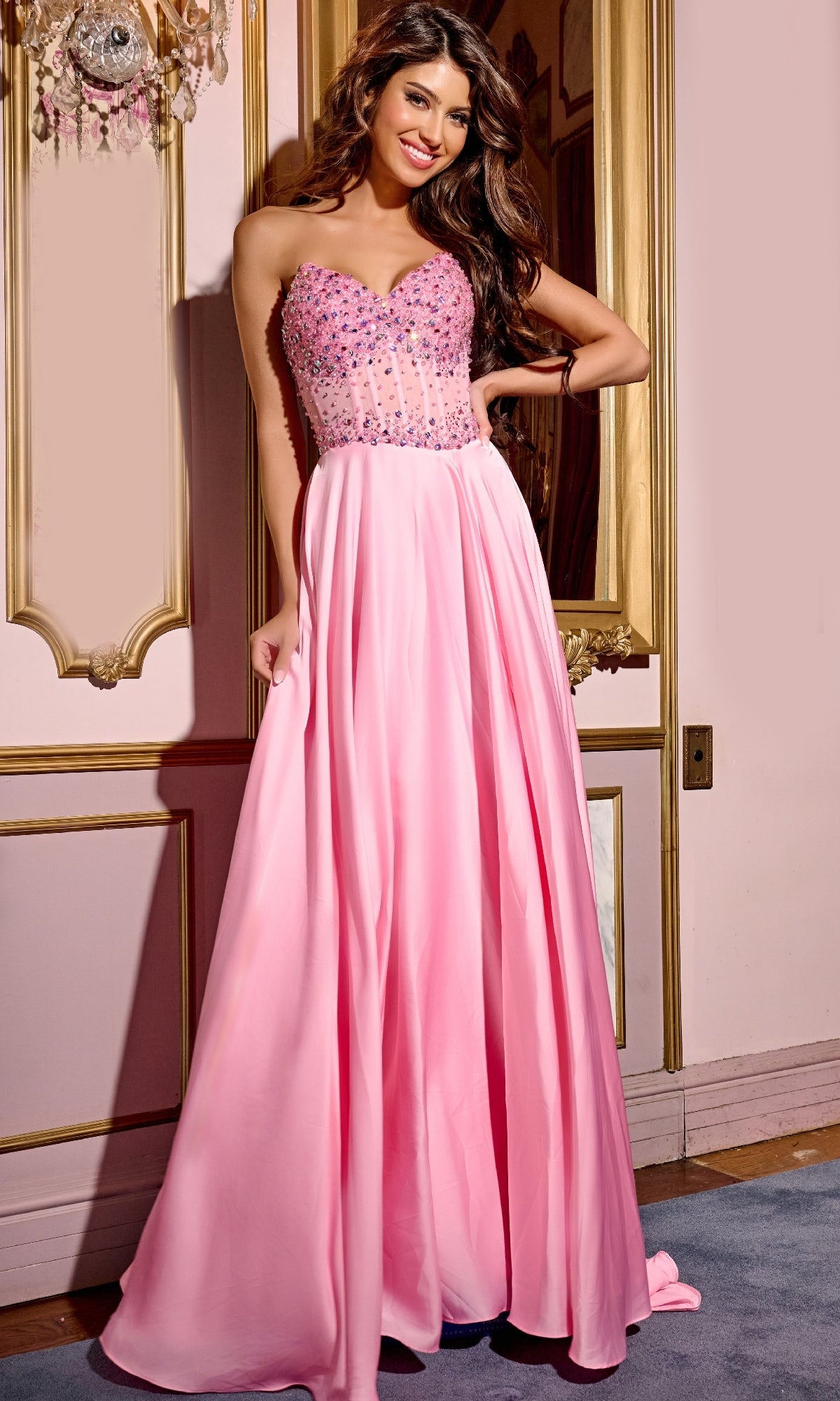 A woman models the Jovani D6021 pink long prom dress, featuring a strapless beaded corset bodice, while standing indoors against an ornate gold-trimmed wall.