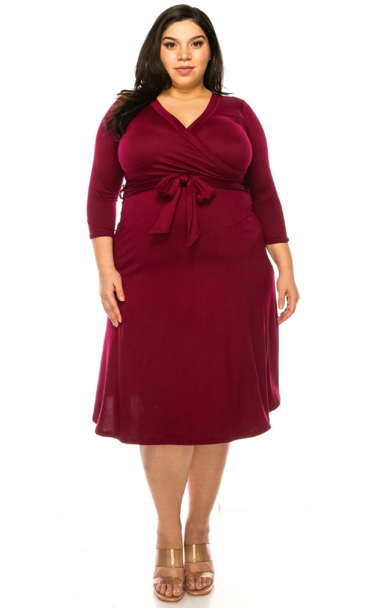 A woman wears the Faux-Wrap Plus-Size Casual Dress with Sleeves in burgundy, featuring a knee-length cut, long sleeves, and a front tie, standing and facing forward against a white background.