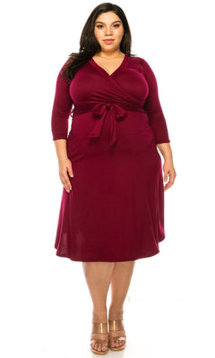 A woman wears the Faux-Wrap Plus-Size Casual Dress with Sleeves in burgundy, featuring a knee-length cut, long sleeves, and a front tie, standing and facing forward against a white background.
