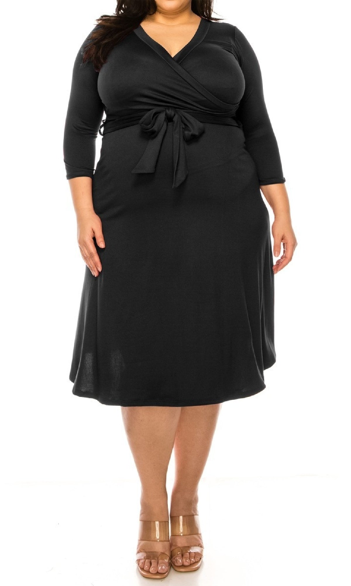 A person wearing the Faux-Wrap Plus-Size Casual Dress with Sleeves in black, featuring three-quarter sleeves and a tied waist, pairs it with tan open-toe sandals while standing against a white background.