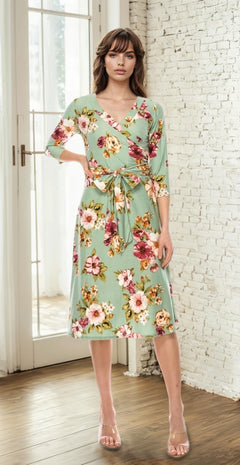 A woman poses indoors with one hand on her hip, wearing the Sage Green Floral Print Faux-Wrap Casual Dress and nude heels, standing in front of a white brick wall and window.