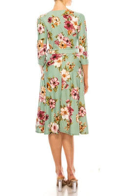 A woman stands with her back to the camera, wearing the Sage Green Floral Print Faux-Wrap Casual Dress with a v-neckline and three-quarter sleeves, paired with clear-heeled shoes.