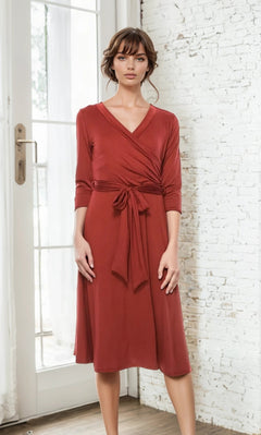 A woman stands indoors in front of a white brick wall and glass door, wearing the Faux-Wrap Casual Short Dress with Sleeves—a knee-length rust dress featuring long sleeves, a tied waist, and a v-neckline.
