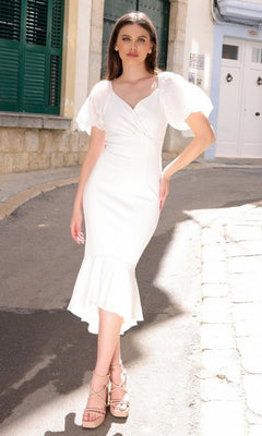 A woman stands on a sunlit street in the Velvi Diana Short Sleeve Midi Semi-Formal Dress—white, fitted, with ruffled hem and puff sleeves. Perfect for parties or as a rehearsal dinner dress.